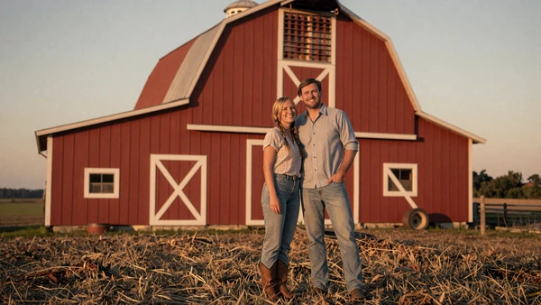 Happy farming couple in front of a red barn at sunset, success stories