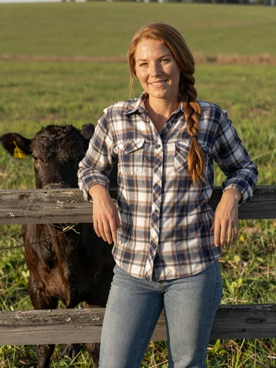 Hannah, 30, Iowa cattle rancher leaning on a wooden fence beside black Angus calves in green pasture