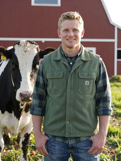 Kyle, 34, Wisconsin dairy farmer smiling beside a Holstein cow in a green pasture with red barn in background